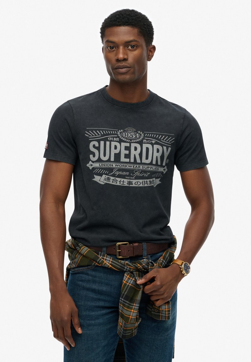 Zwarte katoenen T-shirt met een witte graphic print met de tekst "SUPERDRY". Draagt bij blauwe jeans en een groen geruit overhemd om de taille gewikkeld.