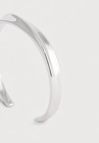 Brățară din argint curbă, lustruită, cu o suprafață netedă și reflectantă, pe un fundal alb simplu.