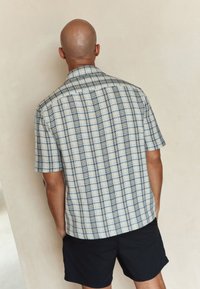 Camicia a maniche corte con un motivo a quadretti testurizzato in crema e blu, con colletto classico e vestibilità comoda. Indossata con pantaloncini neri.