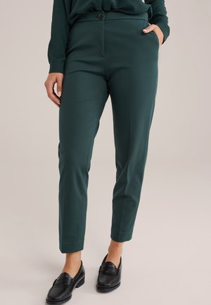 Roheline kitsas sobivusega pükste paar, mis on valmistatud siledast kangast, eesnuppe sulgemisega, küljetaskute ja kitsenevate pahkluude detailidega. Stiliseeritud mustade mokassiinidega.