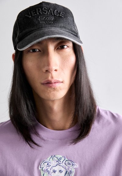 Schwarze Denim-Cap mit erhabenem "Versace"-Logo, getragen mit einem lavendelfarbenen T-Shirt, das ein gesticktes Gesichtsgrafik zeigt. Lange dunkle Haare sind zu sehen.