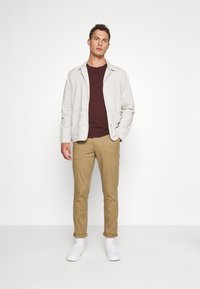 Lichtbeige jasje over een bordeauxrood shirt, gecombineerd met lichtbruine broek en witte sneakers; casual ontwerp met knoopdetails en een relaxte pasvorm.