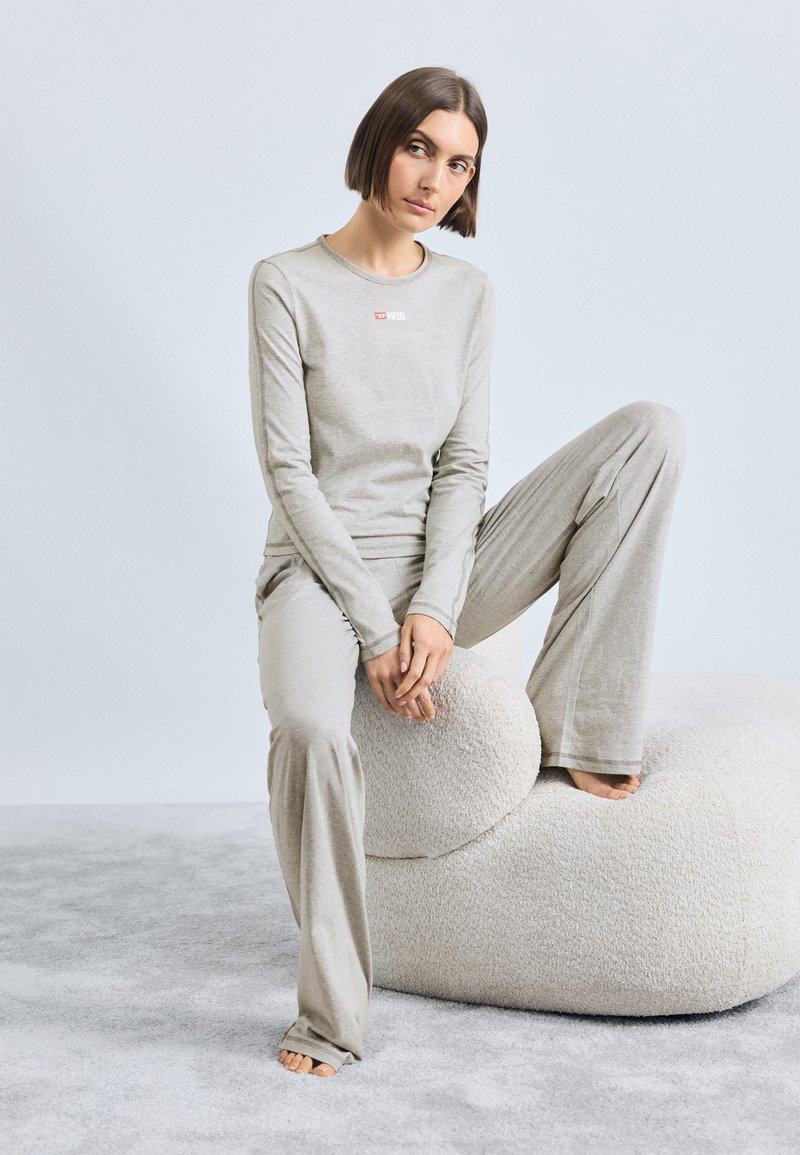 T-shirt de mangas longas cinza com um pequeno logótipo vermelho, combinado com calças largas a condizer, ambos feitos de um tecido suave e texturizado. Modelo sentado em um puff macio.