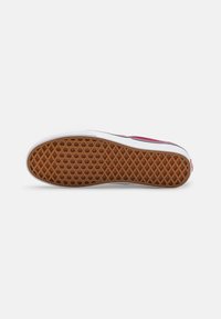 Semelle de chaussure avec motif gaufré brun, semelle intermédiaire blanche et empeigne rouge partiellement visible sur fond blanc.