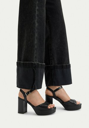 Nogi w czarnych dżinsach o szerokich nogawkach z dużymi mankietami, zestawione z czarnymi sandałami na platformie z odkrytymi palcami, paskiem wokół kostki i grubym obcasem.