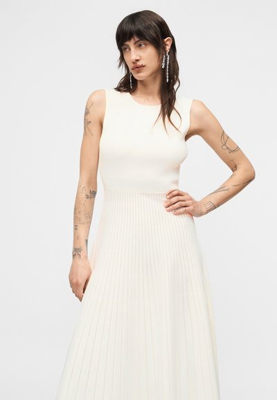 Άτομο με σκούρα μαλλιά, σκουλαρίκια και τατουάζ, φορούν αμάνικο, πλισέ φόρεμα σε κρεμ απόχρωση, στέκεται με το ένα χέρι στη μέση.