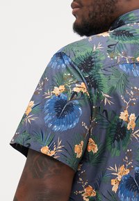 Camisa de manga corta en azul oscuro con hojas verdes y flores naranjas; presenta un vibrante patrón floral, textura suave y diseño de cuello.