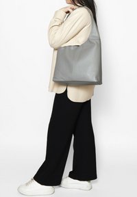 Borsa tote in pelle grigia con una texture morbida, forma triangolare e una sola tracolla, abbinata a un top beige e pantaloni a gamba larga neri.