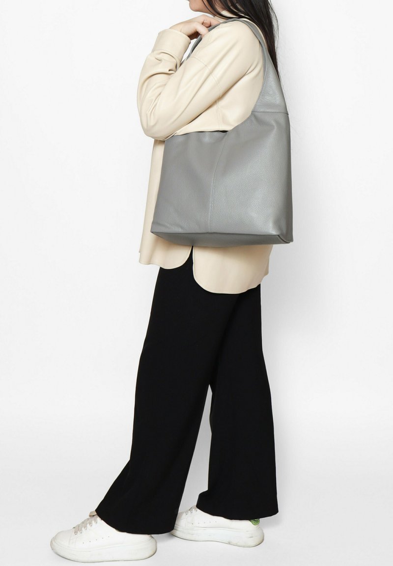 Borsa tote in pelle grigia con una texture morbida, forma triangolare e una sola tracolla, abbinata a un top beige e pantaloni a gamba larga neri.