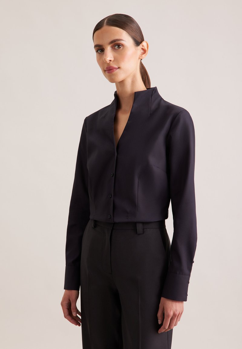 Camicia nera corta con bottoni e collo alto, dalla texture liscia, maniche lunghe e vestibilità su misura, abbinata a pantaloni scuri su misura.