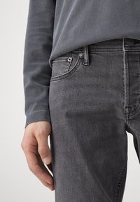 Šedé džíny z denimu s hladkou texturou, přiléhavým střihem a klasickým pětikapsovým provedením. Kombinovány s tmavě šedým dlouhým rukávem.