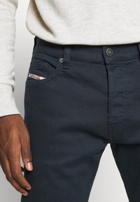 Detalle de unos jeans de un azul marino oscuro con botón de marca y una pequeña etiqueta blanca, combinados con una camiseta de manga larga gris claro, mostrando la mano izquierda relajada.