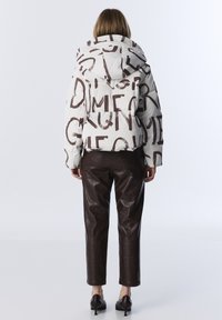 Femme portant une doudoune blanche avec un grand texte marron, un pantalon en cuir marron et des talons hauts marron foncé, se tenant de dos sur un fond blanc.