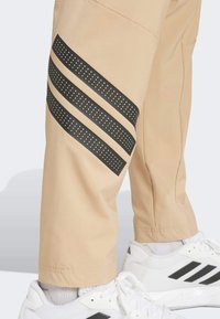 Beige broek met zwarte geperforeerde strepen aan de zijkant. Heeft een ontspannen pasvorm en elastische boorden. Gediend met witte sportschoenen.