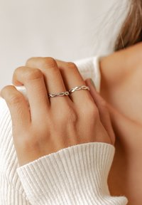 Srebrny pierścionek z przeplatanym wzorem i drobnymi zdobieniami. Dłoń opiera się o fakturowany biały sweter, podkreślając odcień skóry i detale.