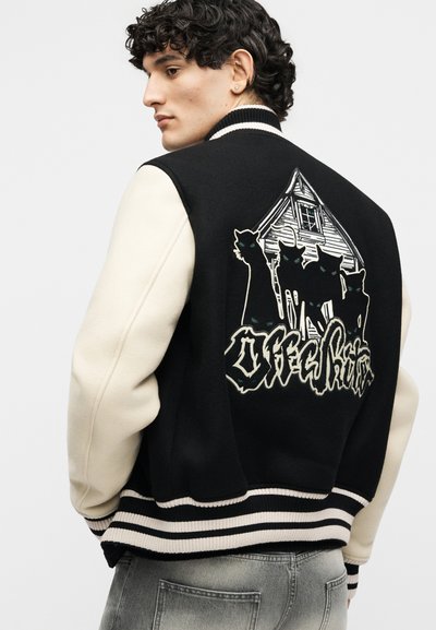 Άντρας που φοράει μαύρο και άσπρο varsity μπουφάν με τέσσερις μαύρες γάτες και ένα σπίτι γραφικό στην πλάτη, συνδυασμένο με γκρι τζιν.