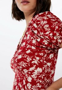 Rotes Blumenkleid mit puffigen Ärmeln, einem taillierten Oberteil und weißen Blumenmustern auf einem strukturierten Stoff. Betonter tiefer Ausschnitt.