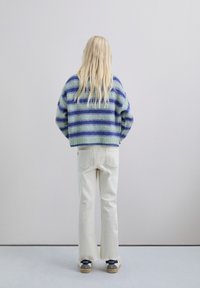 Suéter de lana a rayas en verde y azul, de diseño corto. Jeans de campana beige con dobladillo sin terminar, y zapatos grises con detalles en negro, vistos desde detrás.