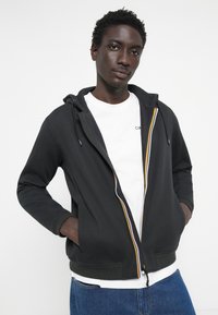 Svart zip-up hettegenser med hette, med flerfarget glidelåsdetalj og ribbestrikkede cuffs. Brystet over en hvit t-skjorte og blå jeans.