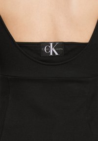 Haut noir sans manches avec un dos échancré, une texture lisse et une étiquette visible portant le logo "Calvin Klein Jeans" en blanc sur fond noir.