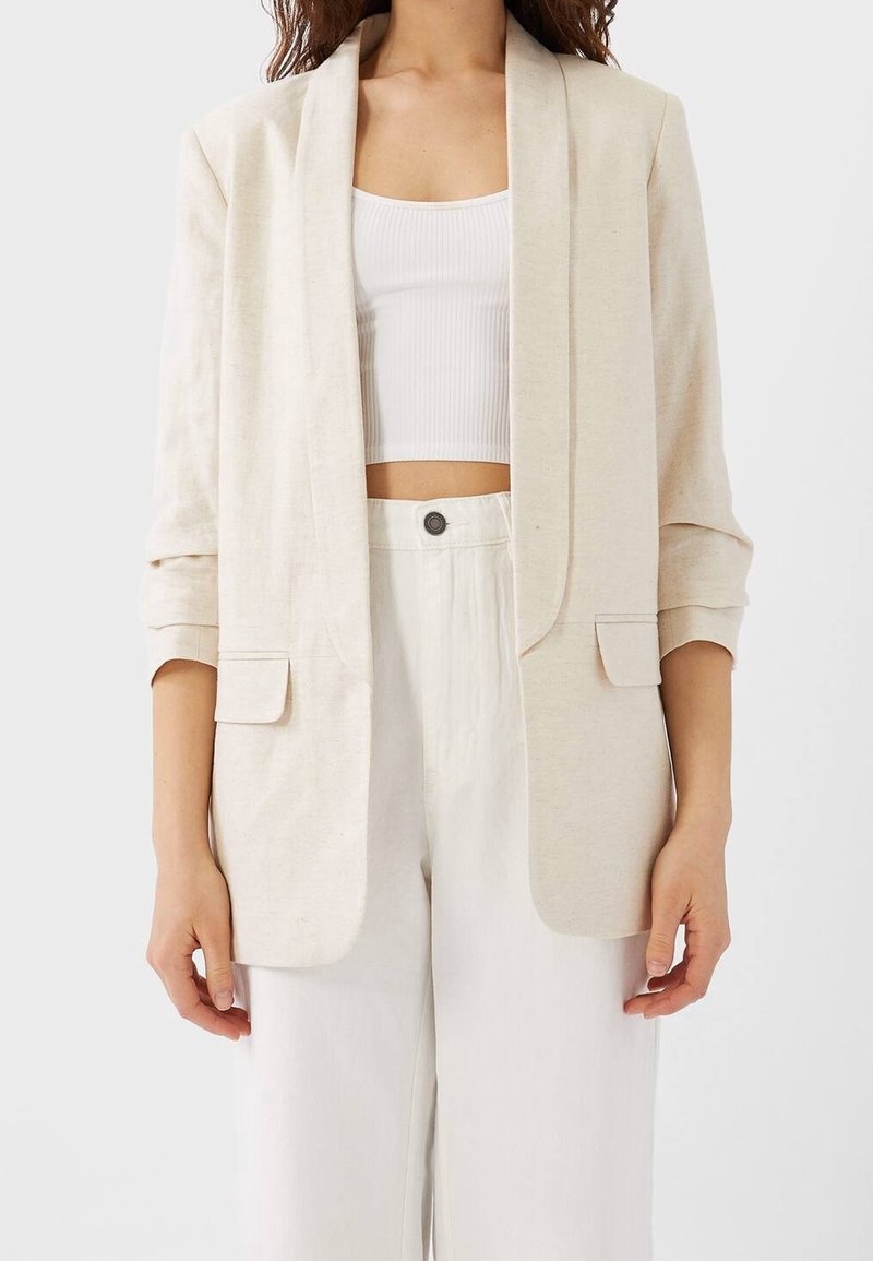 Blazer en lin beige avec un devant ouvert, des revers à crans et deux poches avant. Il présente des manches retroussées et une coupe décontractée.