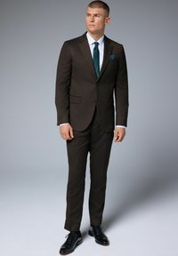 Costume marron sur mesure avec une coupe slim, revers crantés, chemise blanche et cravate vert foncé. Des chaussures en cuir noires complètent le look.