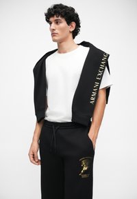 Crna dukserica s logotipom "Armani Exchange" u zlatnoj boji na rukavu, složena preko bijelog majice, u kombinaciji s crnim trenirkama s grafikom.