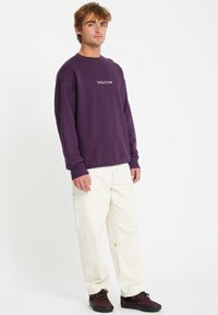 Sweat-shirt violet avec le logo "VOLCOM" blanc, associé à un pantalon blanc et des chaussures foncées. Coupe décontractée avec des poignets côtelés et un tissu texturé.