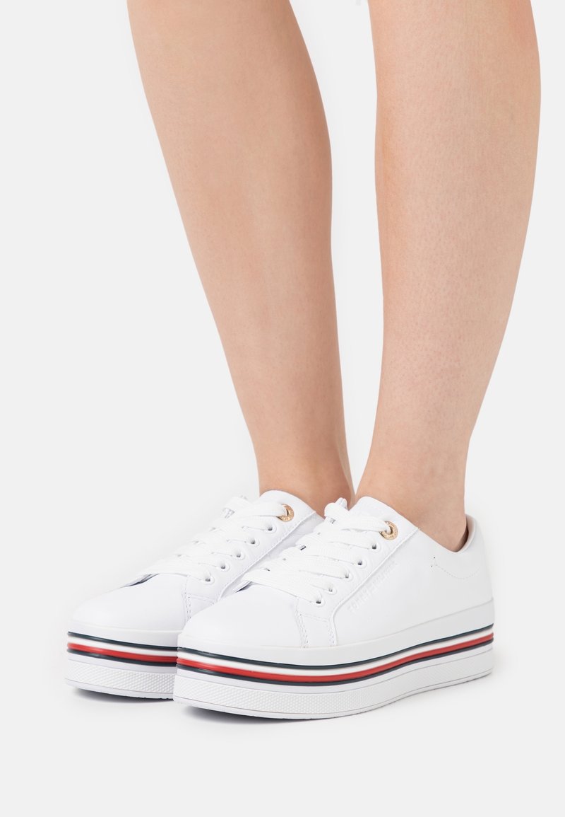 Baskets compensées blanches à texture lisse, avec des œillets dorés et une semelle rayée aux accents rouges et noirs.