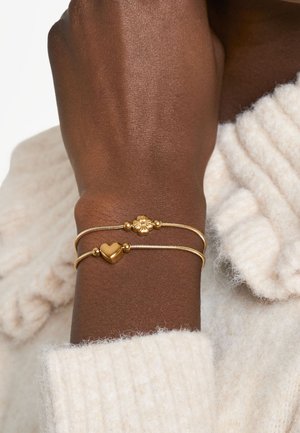 Goldene Armbänder mit Herz- und Blumenanhänger, die glatte, abgerundete Formen und dekorative Perlen aufweisen, getragen an einem Handgelenk mit texturiertem Pullover.