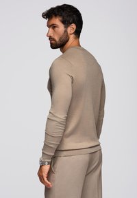 Beige neulepaita, joka on valmistettu pehmeästä neulomateriaalista, ja siinä on pyöreä pääntie sekä pitkät hihat. Istuvuus on lähellä kroppaa, ja hihoissa ja helmassa on joustavat resorit.