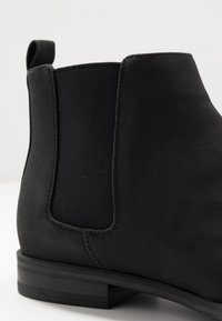 Bota negra de tobillo con suave material de ante, paneles laterales elásticos y un tacón rectangular de goma texturizada. La costura es mínima.