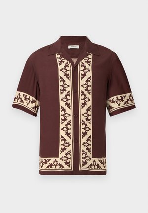 Homme debout les mains dans les poches, portant une chemise marron à manches courtes avec des motifs beige, un pantalon blanc à jambes larges et des chaussures beiges.