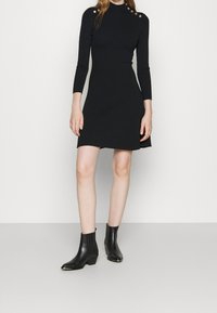 Robe noire à côtes avec un col haut, des manches longues, des accents de boutons dorés sur les épaules et un ourlet évasé. Portée avec des bottines noires.