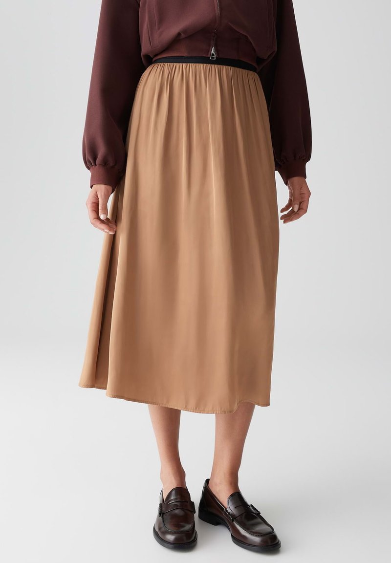 Una gonna midi di colore marrone chiaro, realizzata in tessuto morbido, presenta un design fluido e una cintura elastica, abbinata a mocassini scuri.