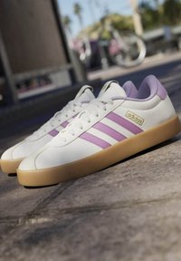 Zapatillas blancas de cuero con rayas moradas, cuello acolchado y suela de goma. Cuenta con una parte superior texturizada y detalle de marca lateral.
