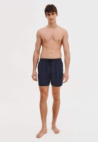 Granatowe kąpielówki z elastycznym pasem, ze sznurkiem i gładką teksturą. Noszone przez stojącego modela mężczyznę bez koszulki.