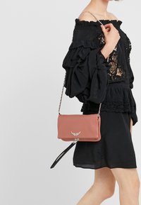 Różowa skórzana torba na ramię z metalowym akcentem i odczepianym czarnym paskiem, o gładkiej teksturze i prostokątnym kształcie.