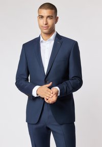 Marineblauer Anzug mit taillierter Passform, ausgestattet mit einem einreihigen Design, braunen Knöpfen und einer schlanken, strukturierten Silhouette. Darunter ein weißes Hemd.