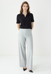 Zwarte polo met korte mouwen gecombineerd met lichtgrijze hooggesneden broek met wijde pijpen en zwarte puntige hakken tegen een neutrale achtergrond.