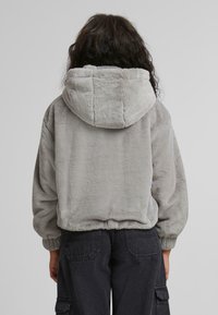 Grå, kort jakke i blødt, fyldigt stof. Har hætte og elastiske ærmer, med et minimalistisk design og ingen synlige mønstre eller metaldele.