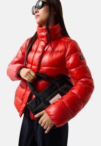 Doudoune rouge avec finition brillante, col haut et poches zippées, associée à un sac noir comportant une étiquette de marque rectangulaire.