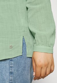 Blusa verde chiaro con texture e orlo spaccato, caratterizzata da un dettaglio logo argento a contrasto vicino al fondo. Abbinata a jeans blu.