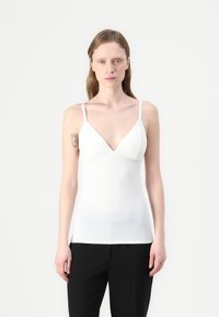White sleeveless top with a v-neck design, soft texture, and fitted silhouette, paired with black pants. Minimalist style and clean lines.