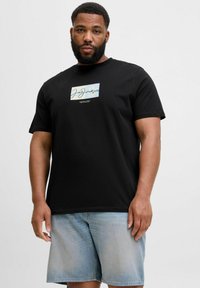 Sort bomulds t-shirt med et rektangulært logo-patch i lyseblå med hvid skrift. Sammen med lyse denimshorts.