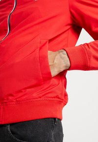 Giacca bomber rossa realizzata in tessuto liscio, con chiusura a zip e polsini a costine. Sul lato sinistro presenta una tasca, con un dettaglio in vita elastica.