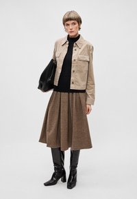 Veste en cord beige, col roulé noir, jupe midi plissée marron et bottes noires montantes élégantes. Sac à main noir inclus.