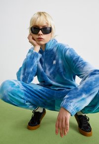 Blå tie-dye fleece joggedrakt med kvart-zip krage, med vide ermer og elastiske mansjetter, parret med svarte sko på en grønn overflate.