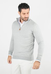 Pull gris clair avec un col zippé, des poignets et un ourlet côtelés. Comprend un accent en cuir sur la fermeture éclair et est porté par-dessus une chemise blanche.