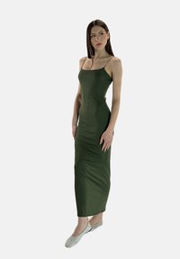 Abito maxi olive green con spalline sottili e silhouette aderente, realizzato in tessuto liscio; modella che indossa scarpe basse decorate.